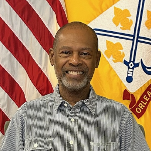 Smiling person with a beard and striped shirt against US and yellow crest flags.